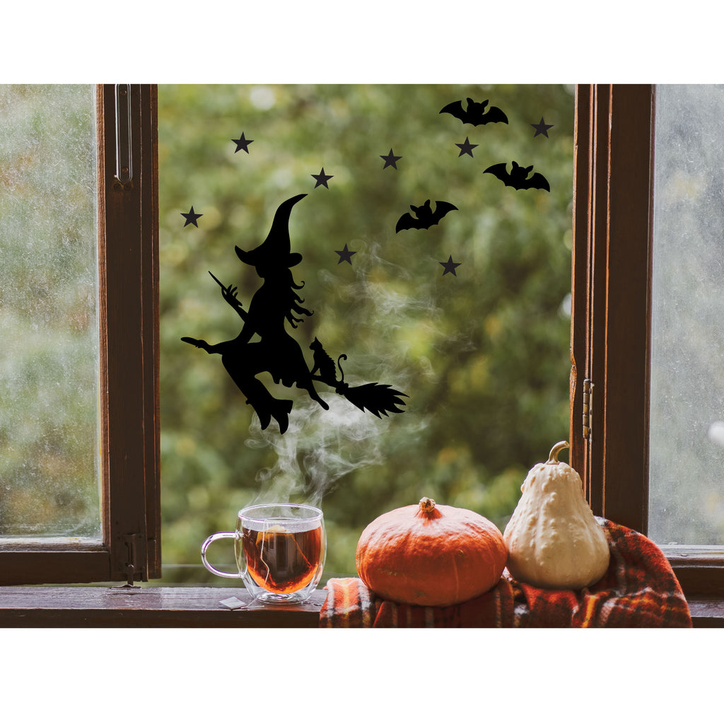 Halloween-themed window decal of a witch flying on a broomstick with pumpkins and tea on a windowsill.