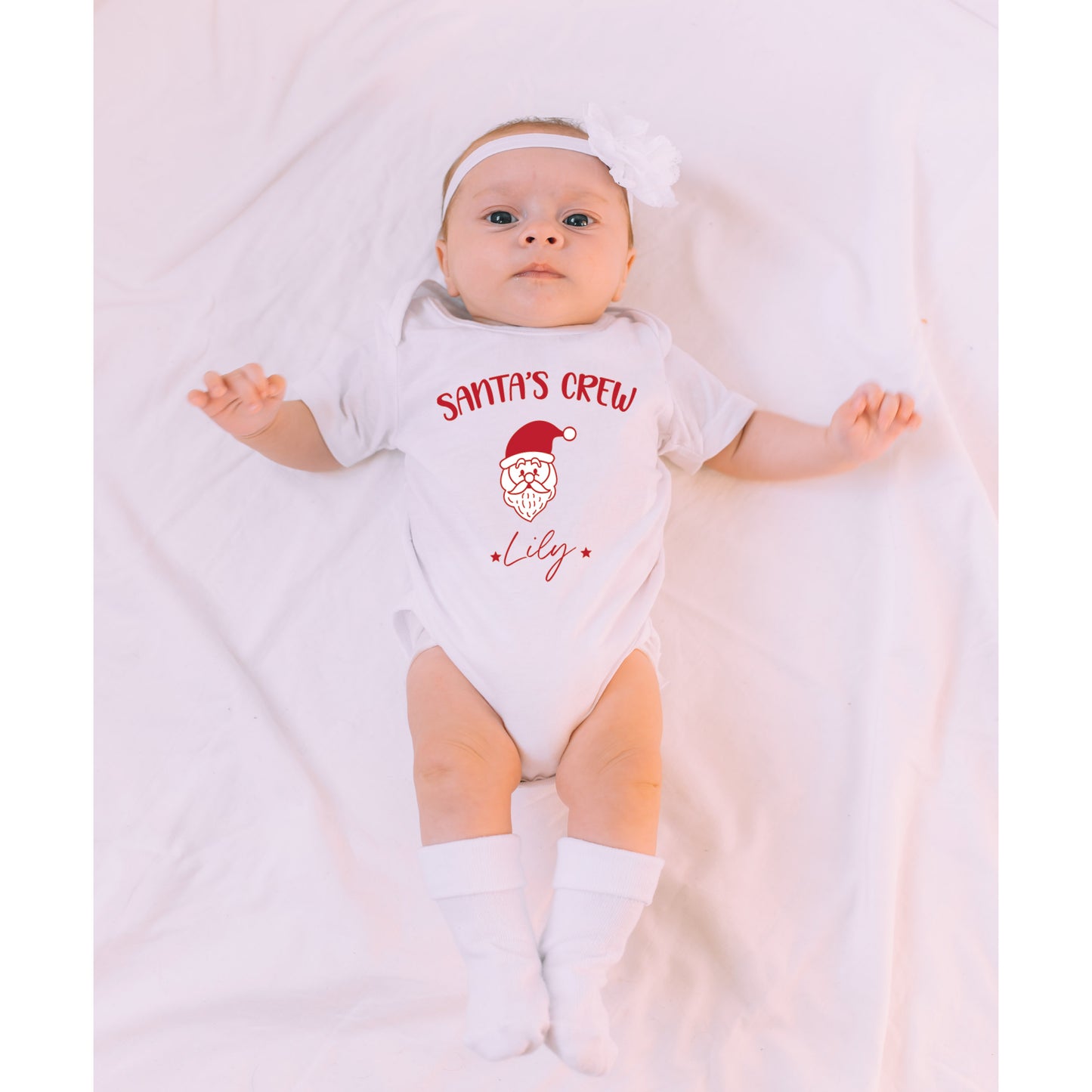 Baby wearing a white onesie with 'Santa's Crew' text and a red beard graphic, lying on a white blanket.