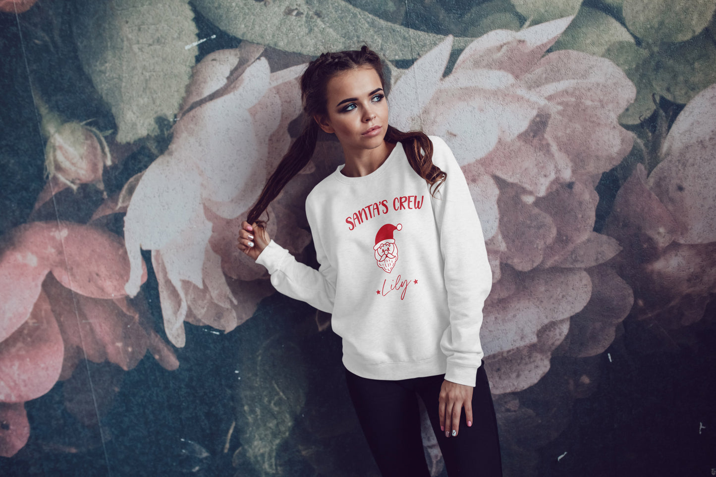 Woman wearing a white sweatshirt with Santa Crew text and graphics, standing against a floral background
